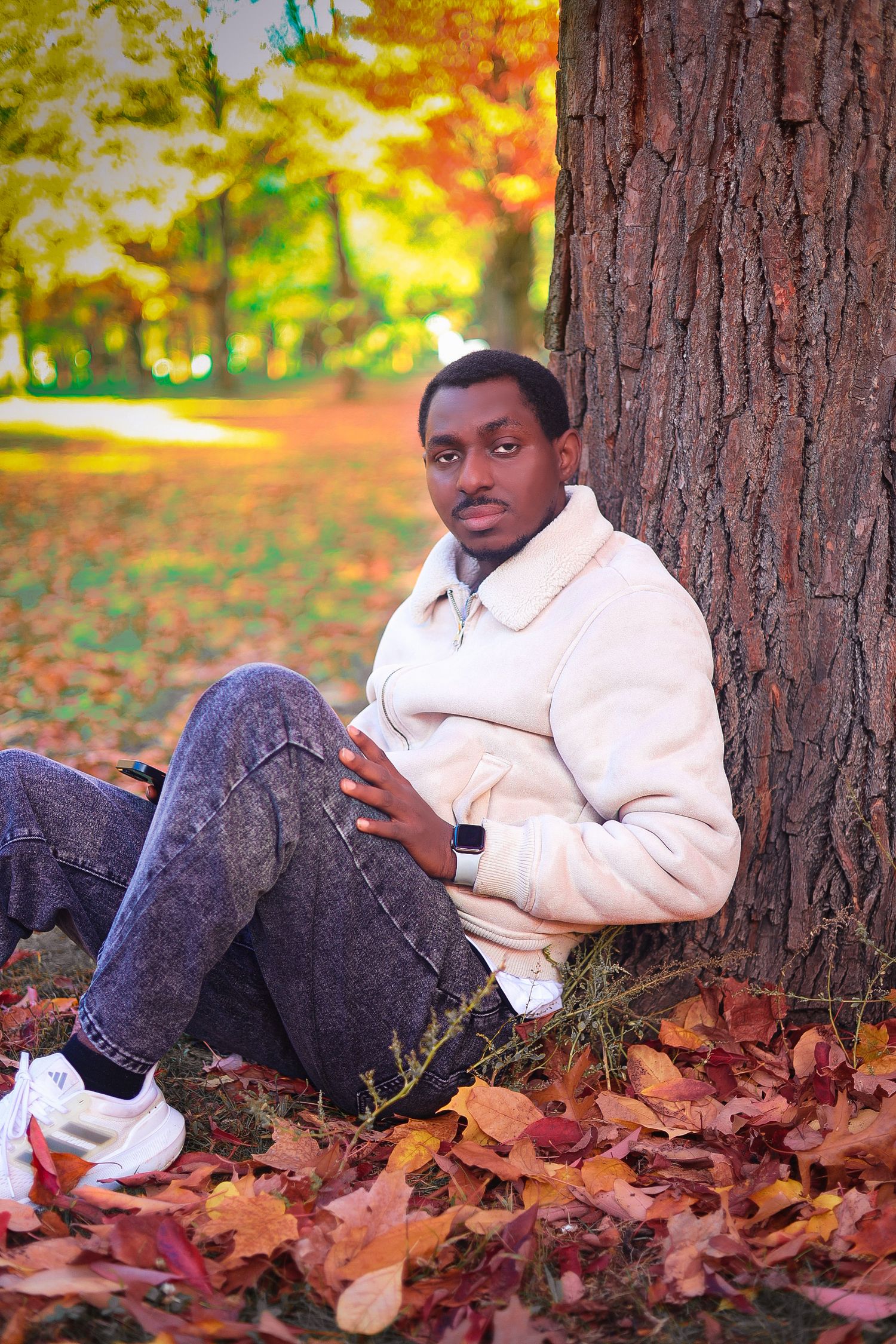 Young Black man in cream sherpa jacket sitting against a tree trunk surrounded by colorful autumn leaves.