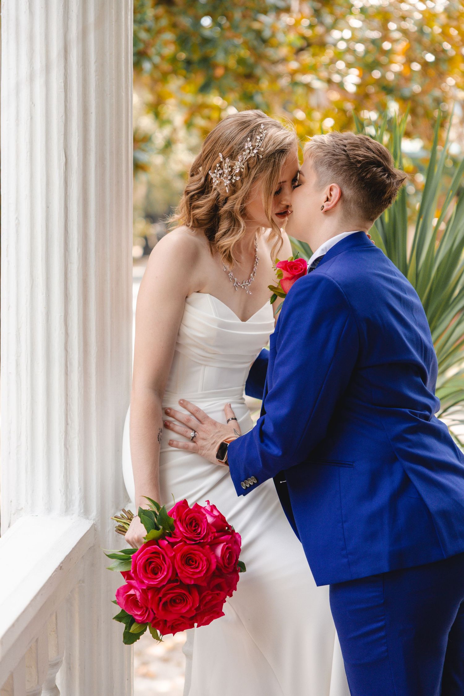 A couple poses for their wedding photo under a tree branch against a white wall with columns.