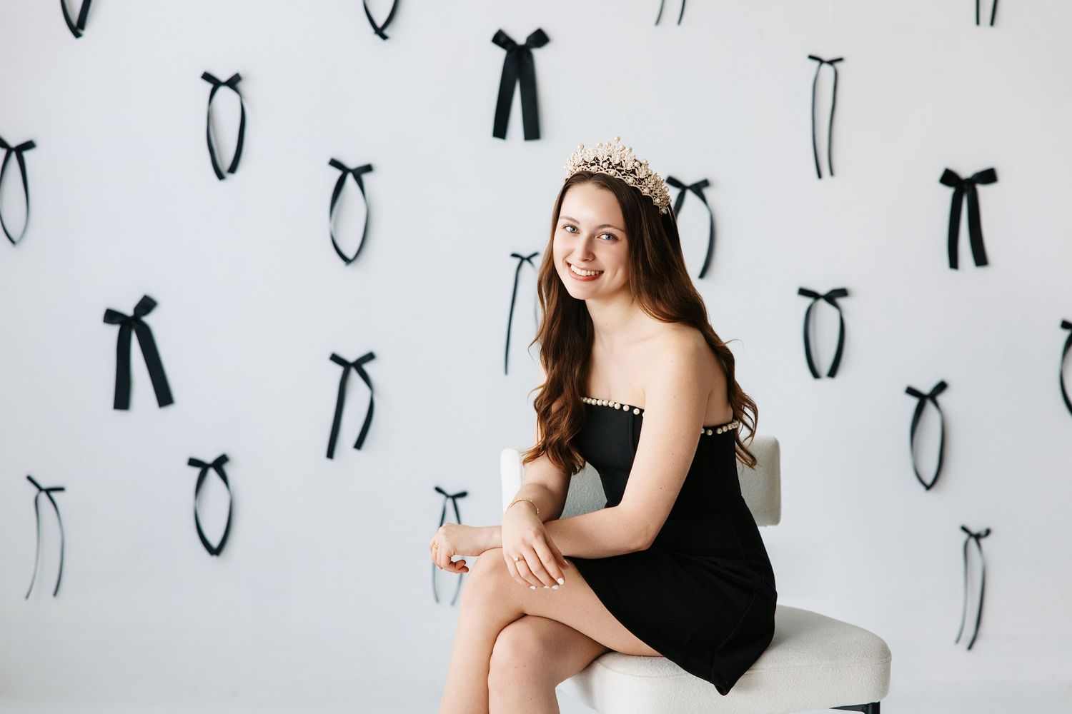A figure in a black dress sits against a white wall decorated with black bow patterns.