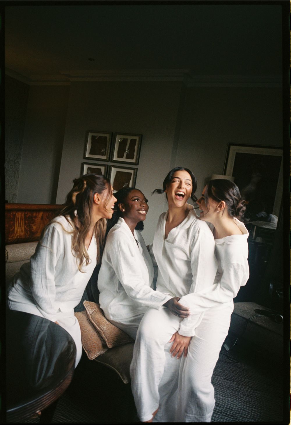Bridesmaids in white robes share laughter in a dimly lit room during wedding preparations.