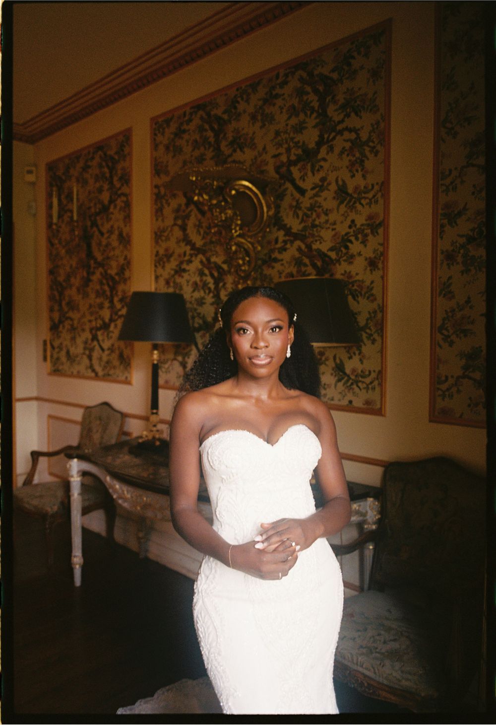 A stunning bride poses in a fitted white strapless wedding gown against an elegant wallpapered backdrop.