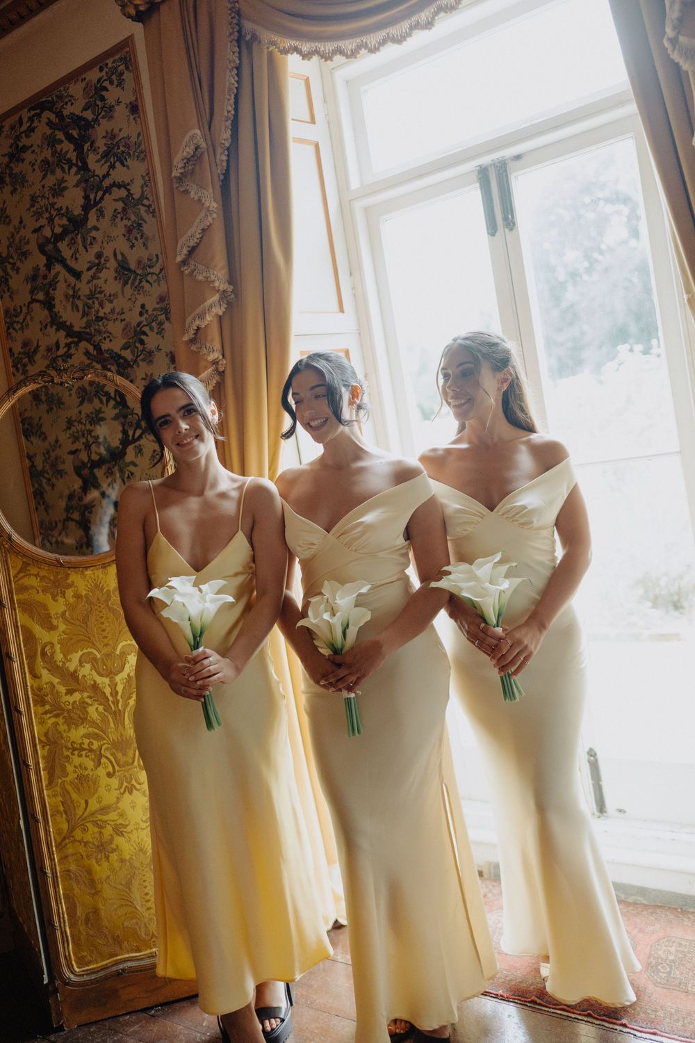 Bridesmaids in cream colored dresses hold white bouquets while posing by window.