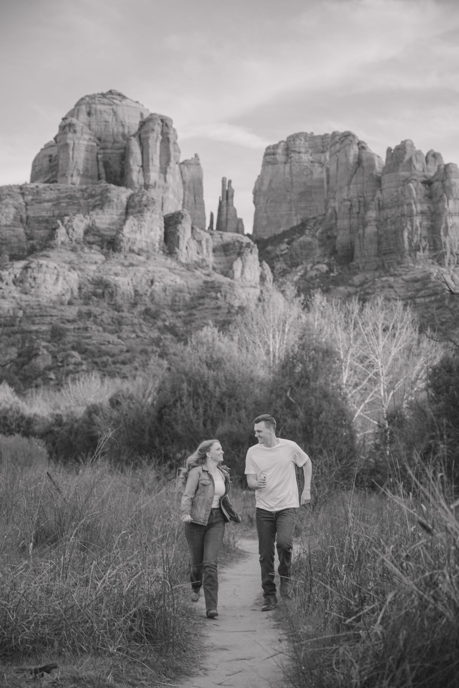 Black and white image of two people walking along a desert trail with towering cliffs behind them.