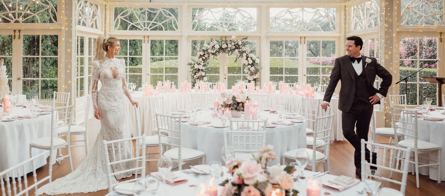 Elegant wedding reception in a sunlit conservatory with floral arch, white tables, and bride and groom standing apart.