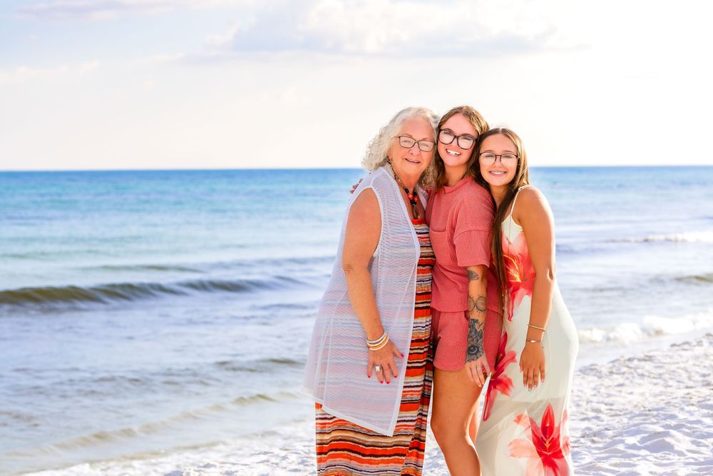 A group in coral and white outfits poses together on a sunny beach with ocean waves in the background.