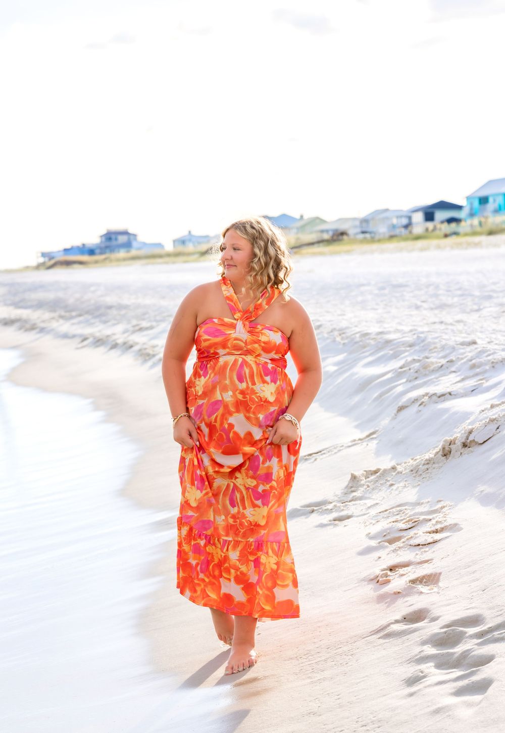 A person in a vibrant orange floral dress poses along a sunny beach shoreline with waves and sand.