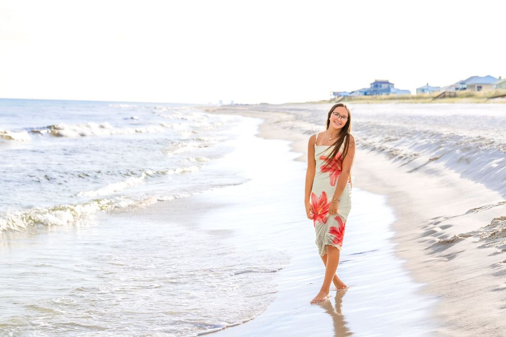 Person in a vibrant floral dress walks barefoot along the shoreline as waves gently lap at the sandy beach.