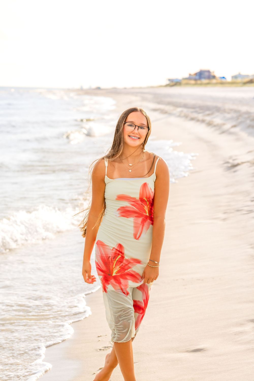 Woman in coral floral top and khaki shorts walks along sandy beach shoreline at sunset.