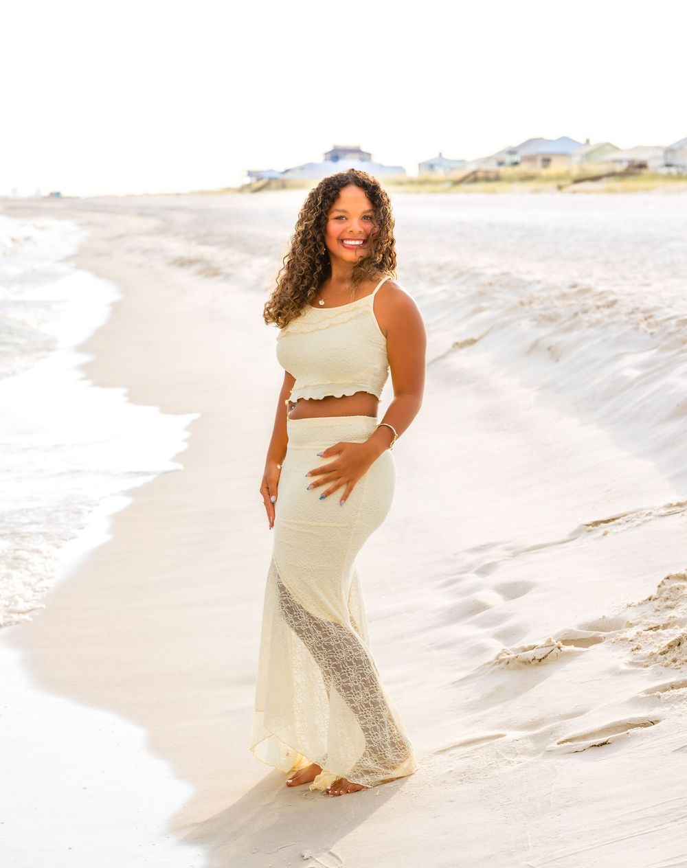 A person in a white dress poses gracefully on a sandy beach with waves in the background during a photoshoot.