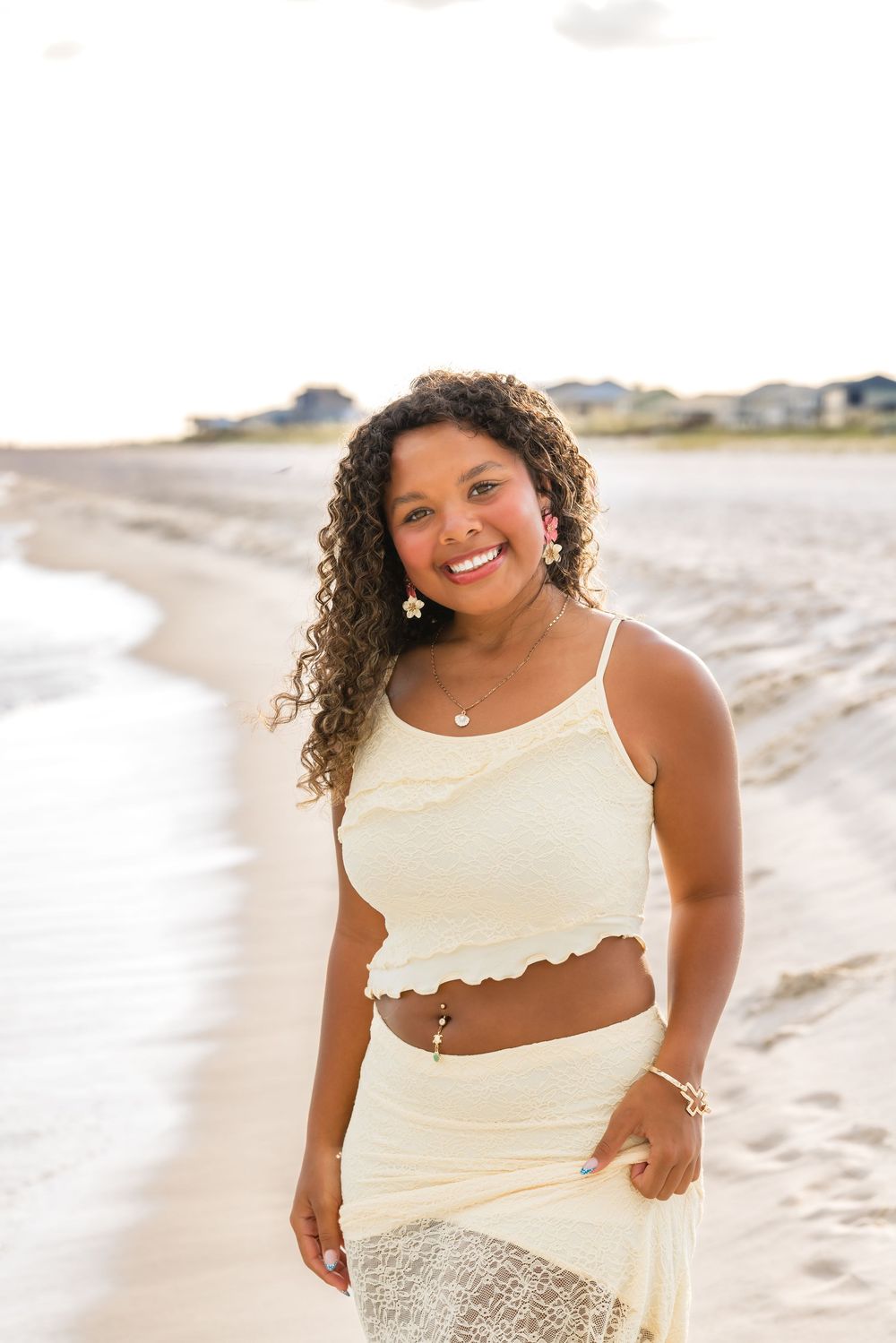 A person with curly hair wearing a cream-colored two-piece outfit poses while smiling on a sunny beach.