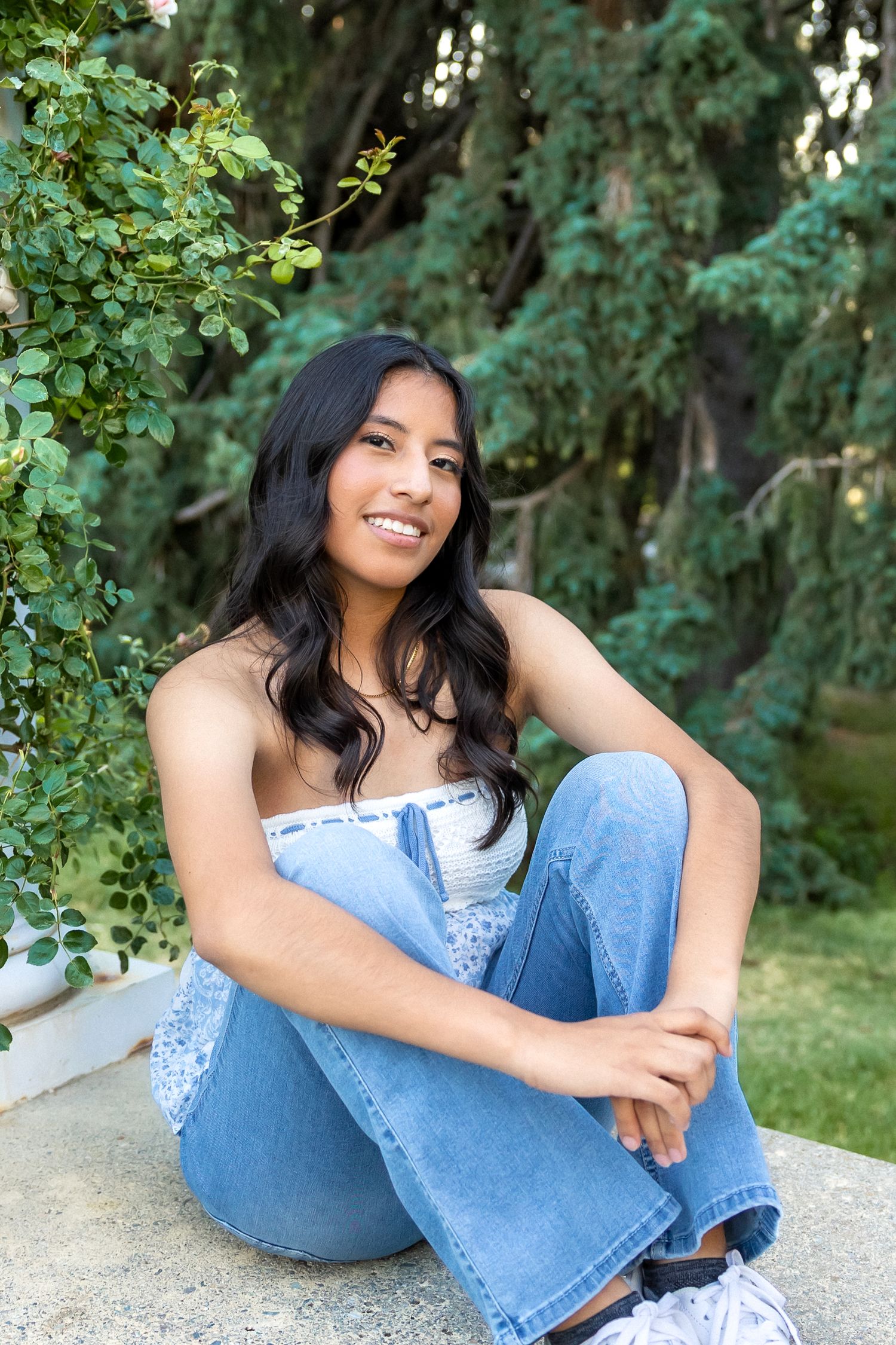 A person in a blue top and jeans sits casually on the ground in a lush garden setting with green foliage in the background.