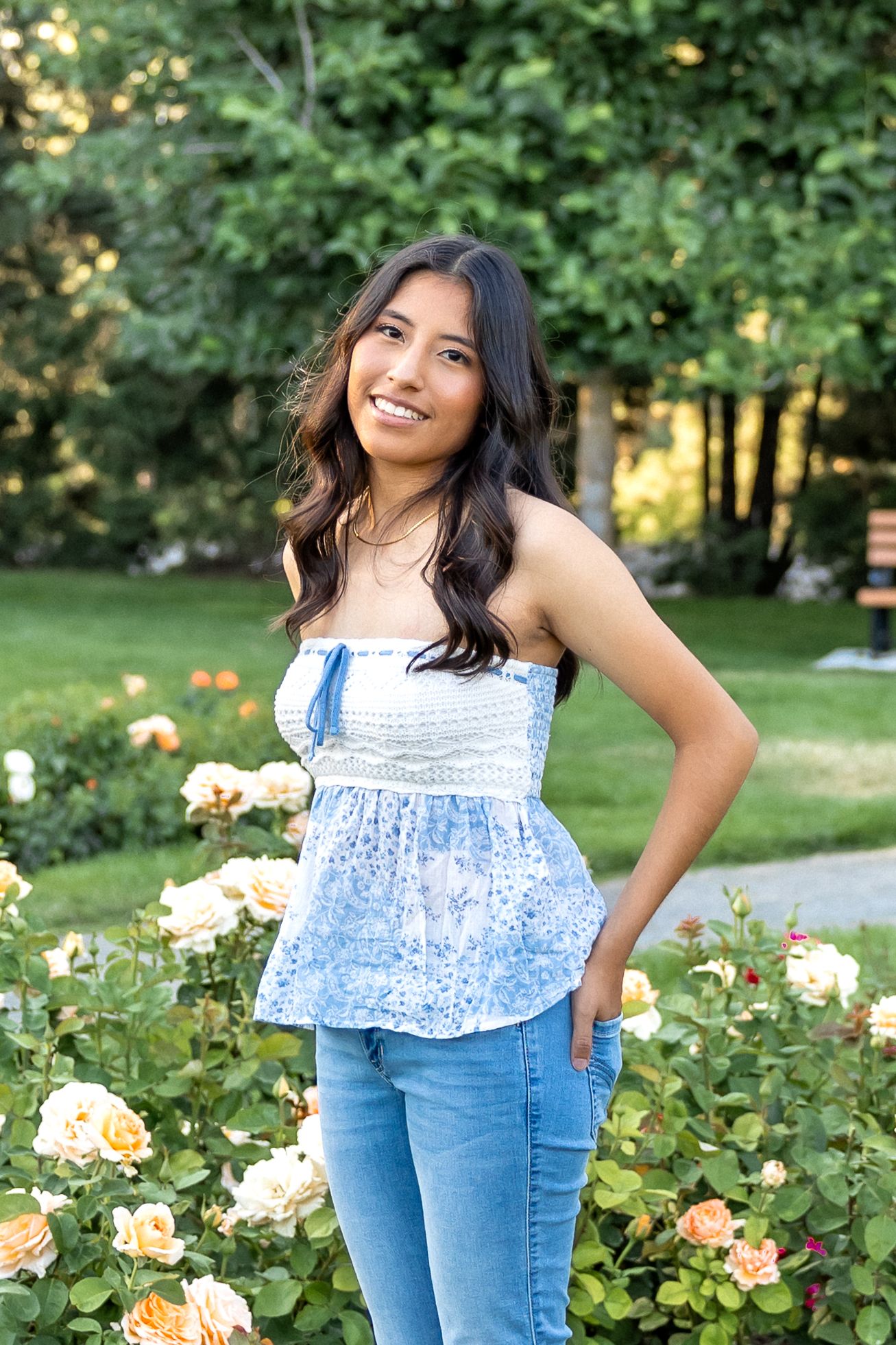 A person in a blue floral top and jeans poses in a garden with blooming roses on a sunny summer day.