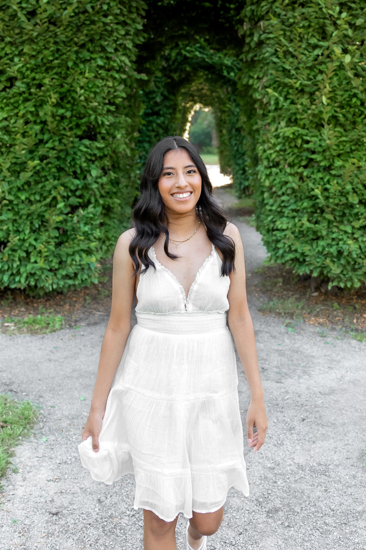 A person in a flowing white summer dress poses outdoors against a green garden backdrop.