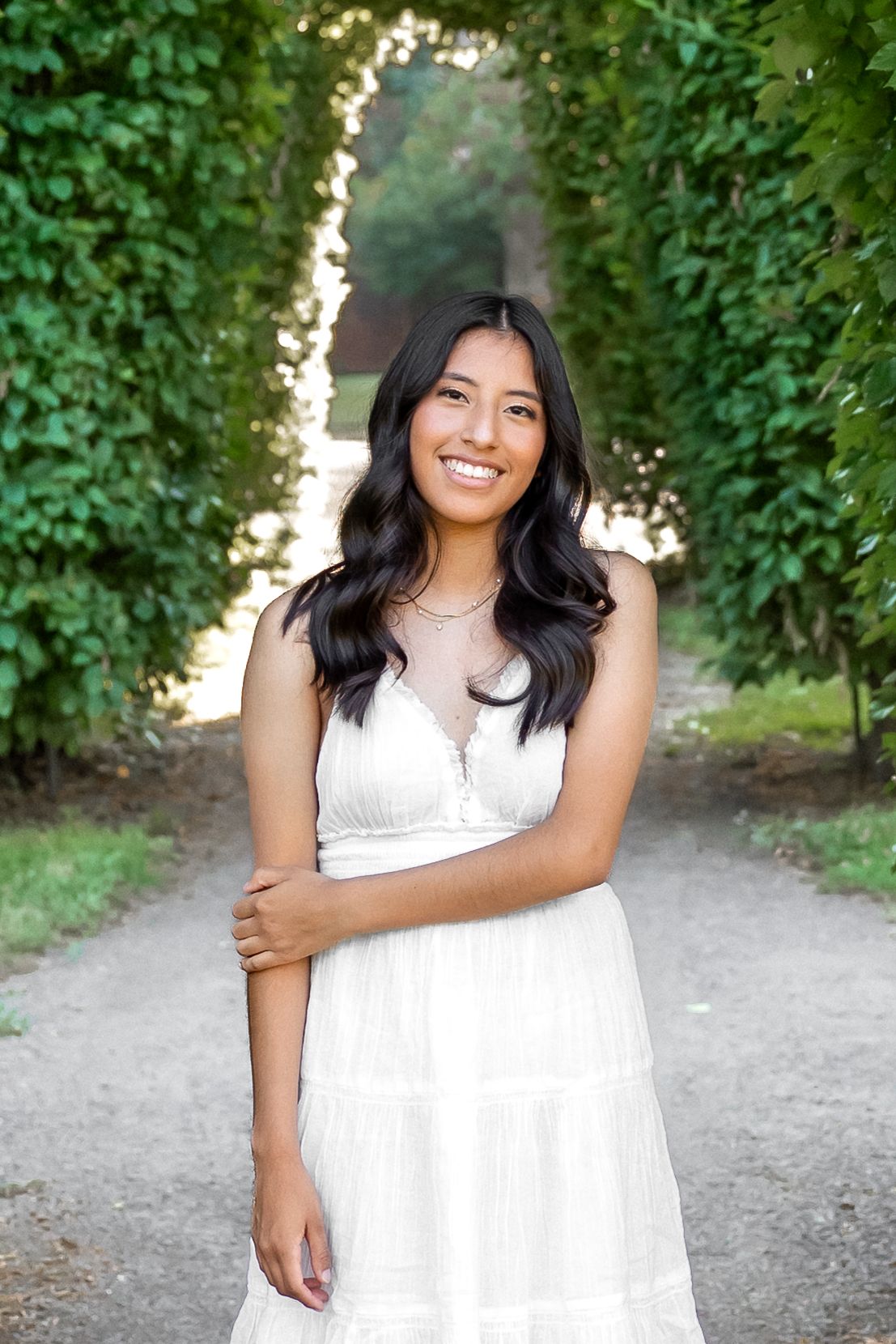 A smiling person in a white dress stands on a path surrounded by lush green foliage on a sunny day.