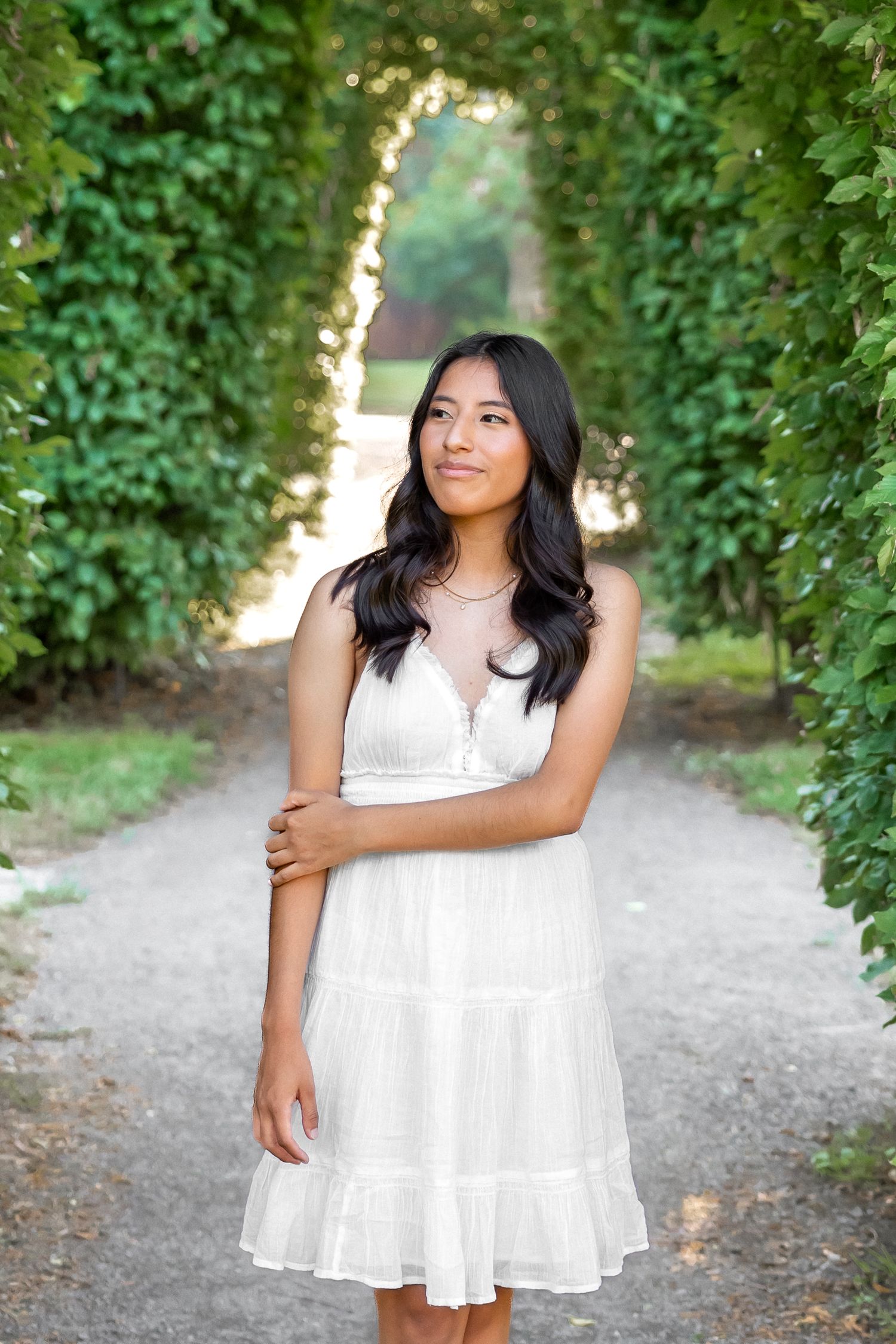 A person in a flowing white dress poses along a tree-lined path on a sunny summer day.