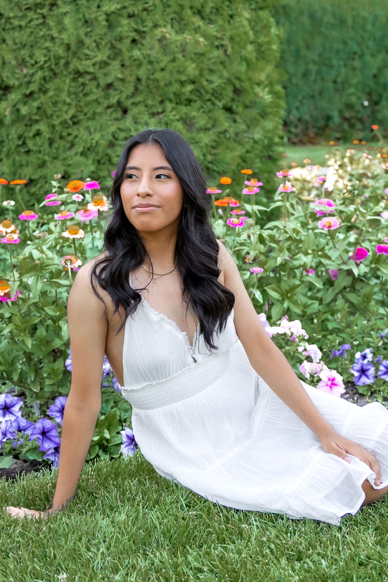 A person in a white summer dress sits gracefully on grass surrounded by colorful garden flowers.