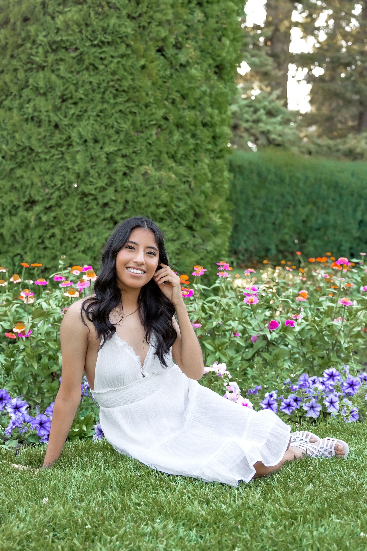 A person in a white dress poses gracefully on grass surrounded by colorful purple and pink flowers in a garden setting.