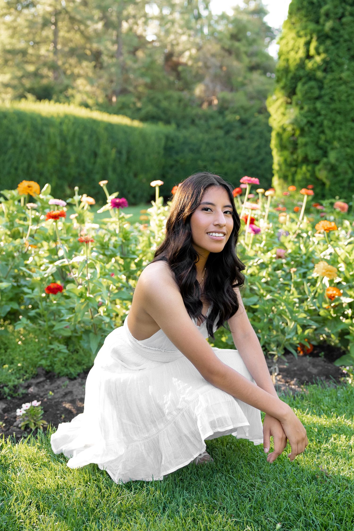 A person in a white dress lies gracefully on green grass surrounded by colorful garden flowers on a sunny day.