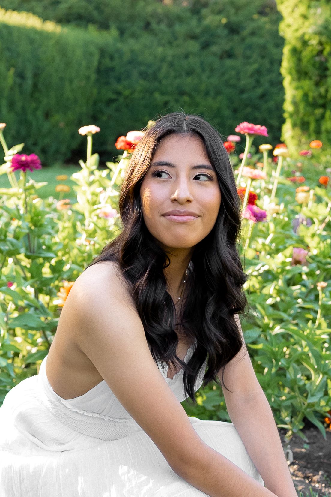 A person in a white dress sits in a garden with colorful zinnias and hedges in the background.