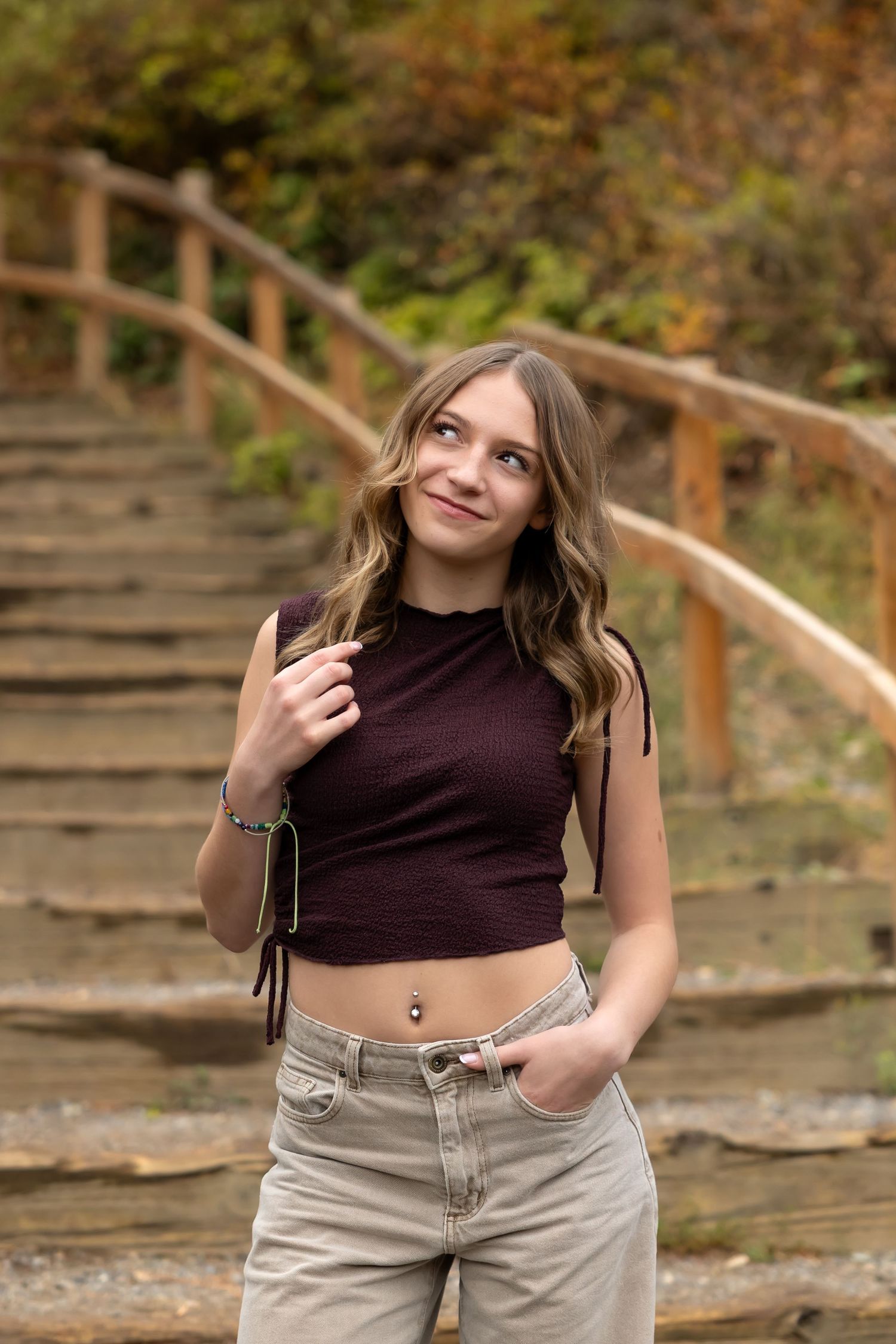 A person in a burgundy crop top and khaki pants poses on stone steps with wooden railings in an autumn setting.