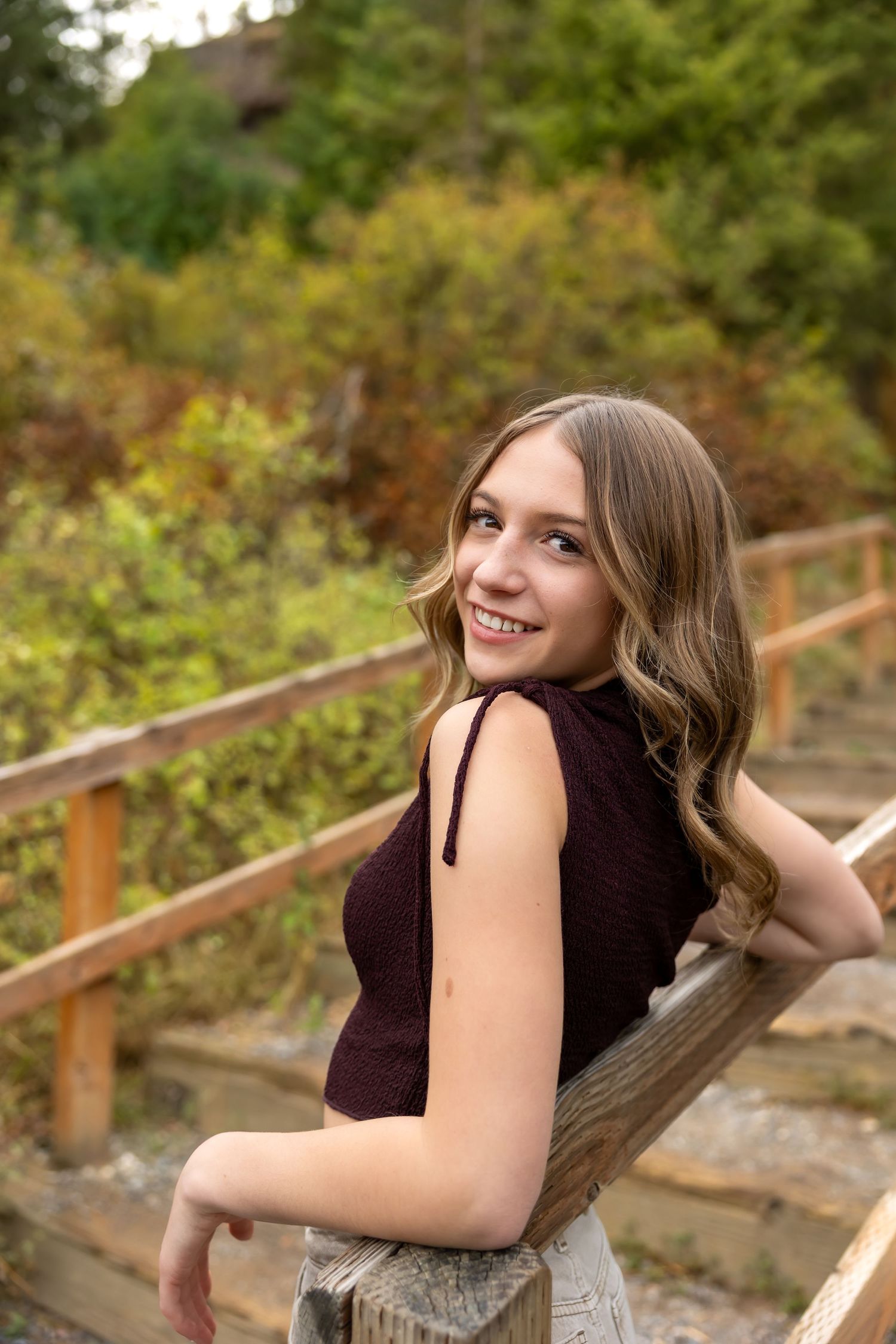 A person in a dark dress poses outdoors on wooden stairs surrounded by lush greenery in a natural setting.