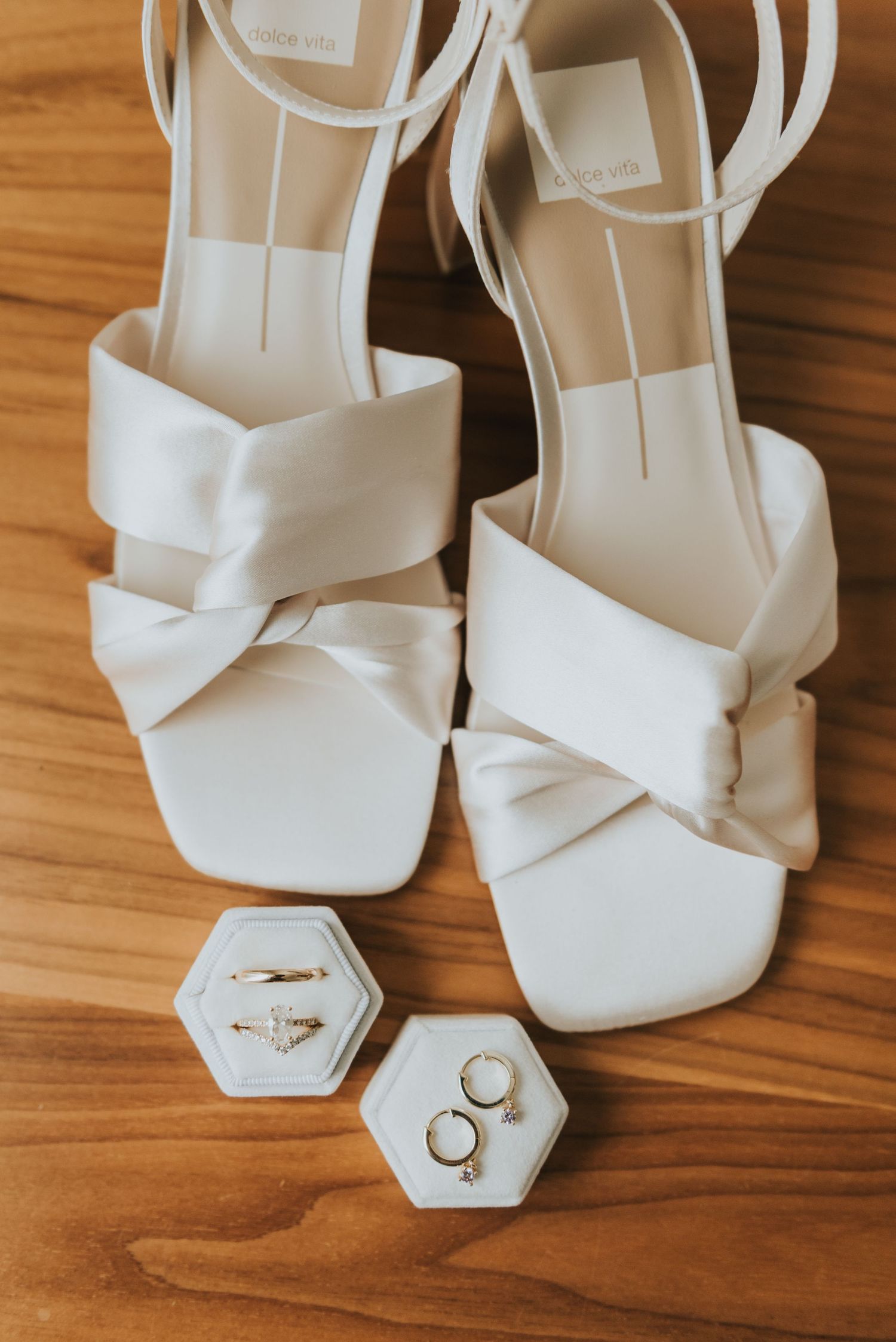 White bridal shoes with bow details and wedding rings displayed on a wooden surface.