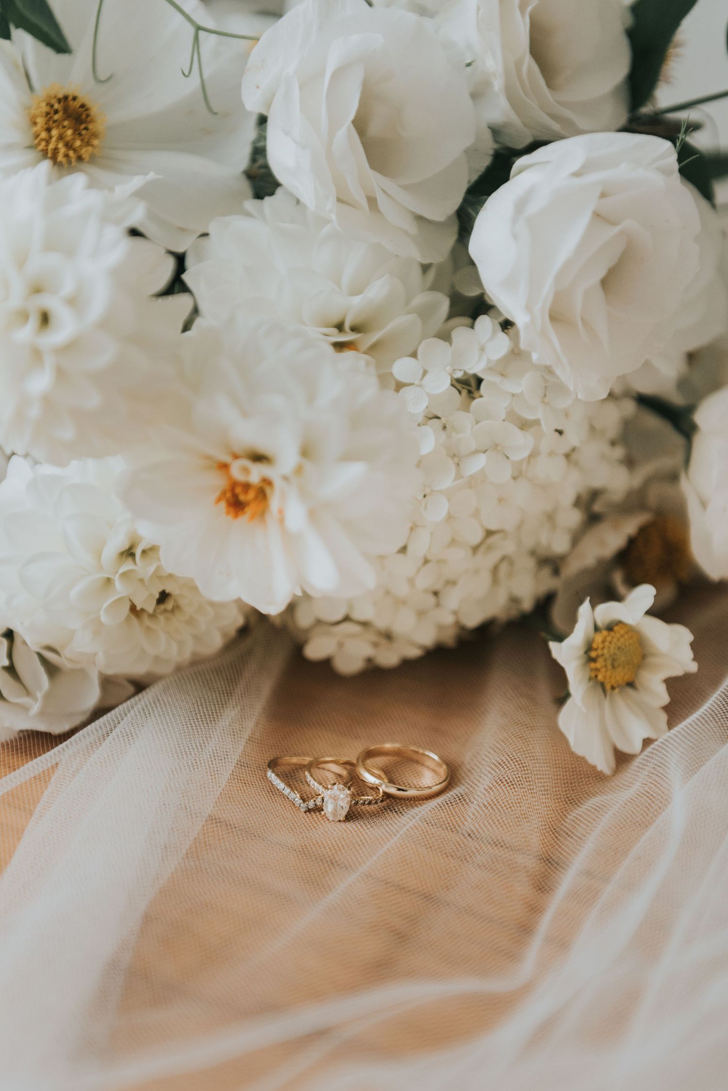 Two gold wedding rings rest beside white roses and delicate floral arrangements on a soft beige surface.