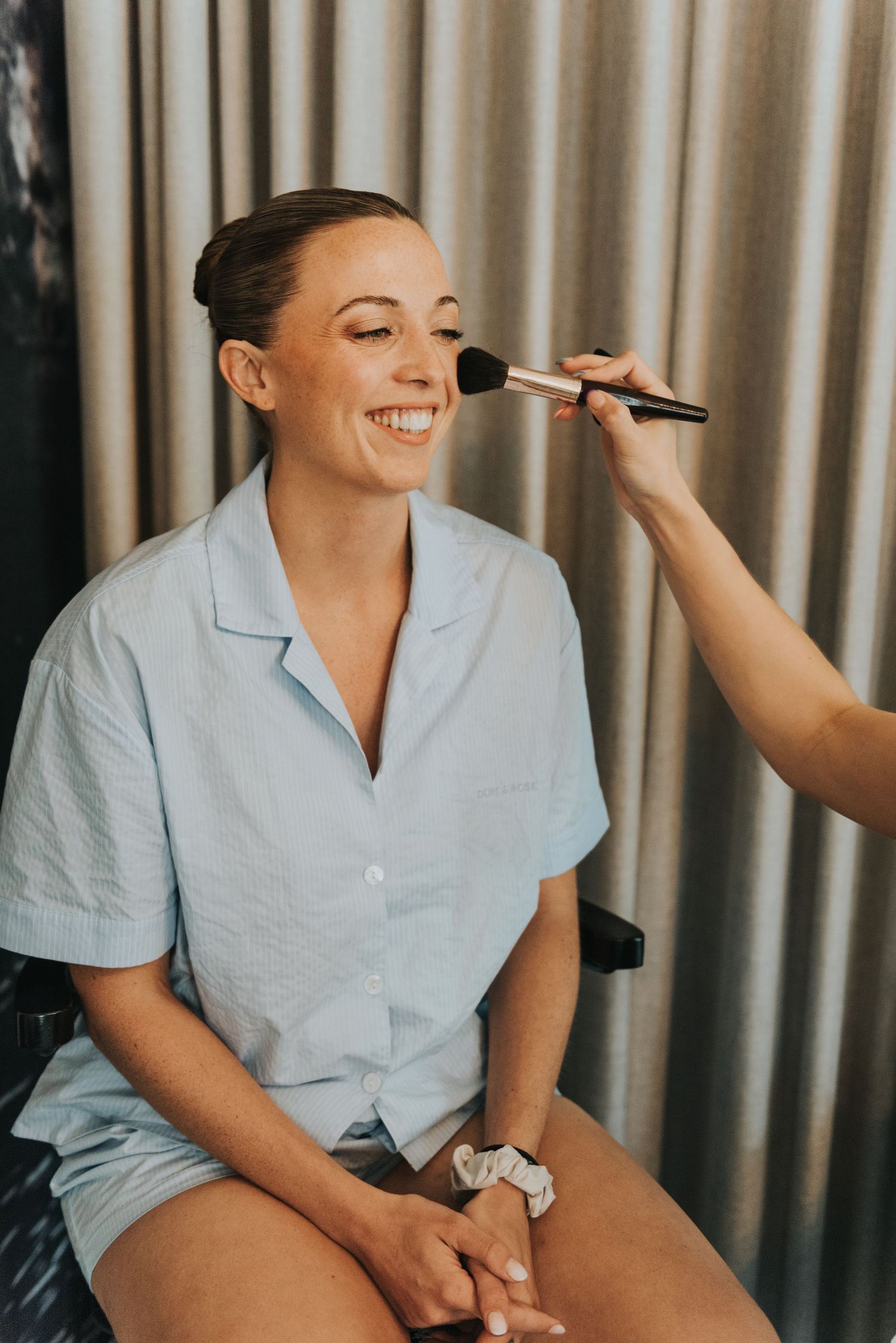 Professional makeup artist applies cosmetics to smiling client wearing light blue shirt in studio setting.