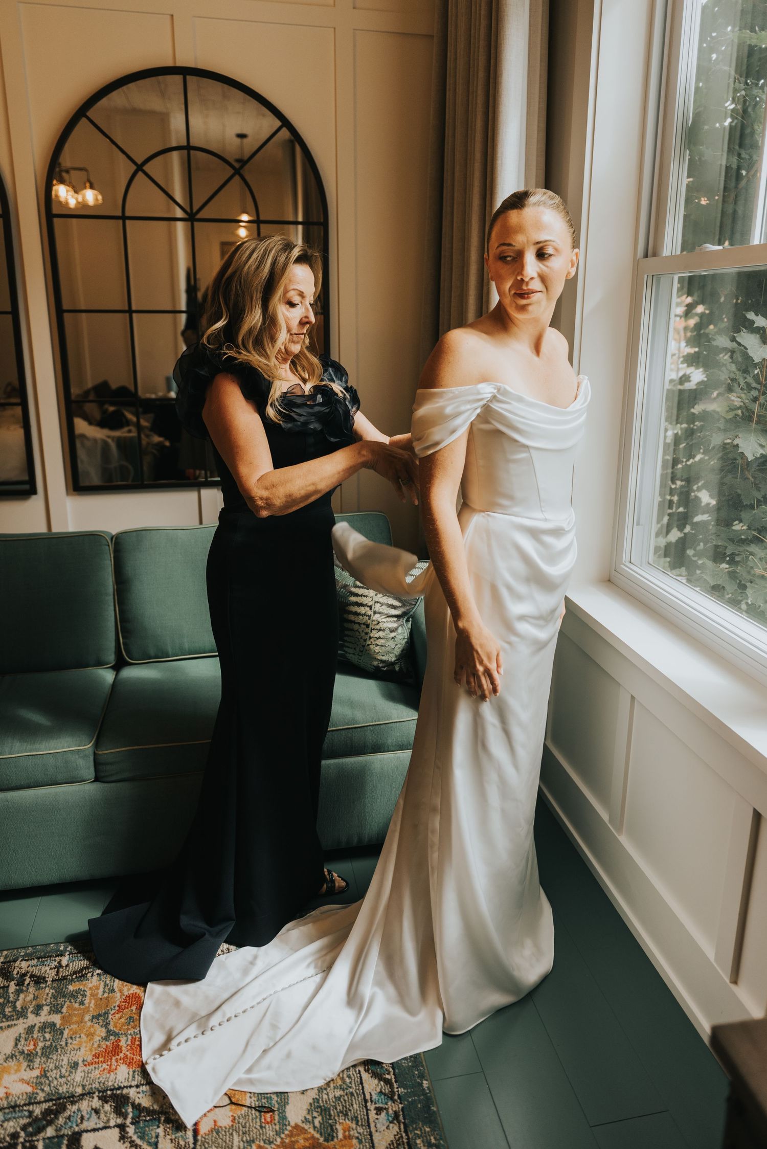 Two women in elegant dresses stand by a window, one helping adjust the other's white wedding gown.
