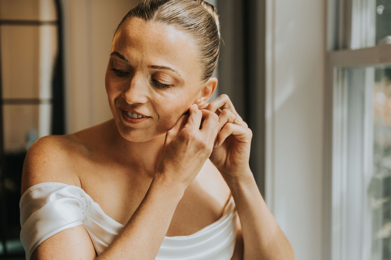 Woman in white dress smiling and touching her earring in a warm-lit room.