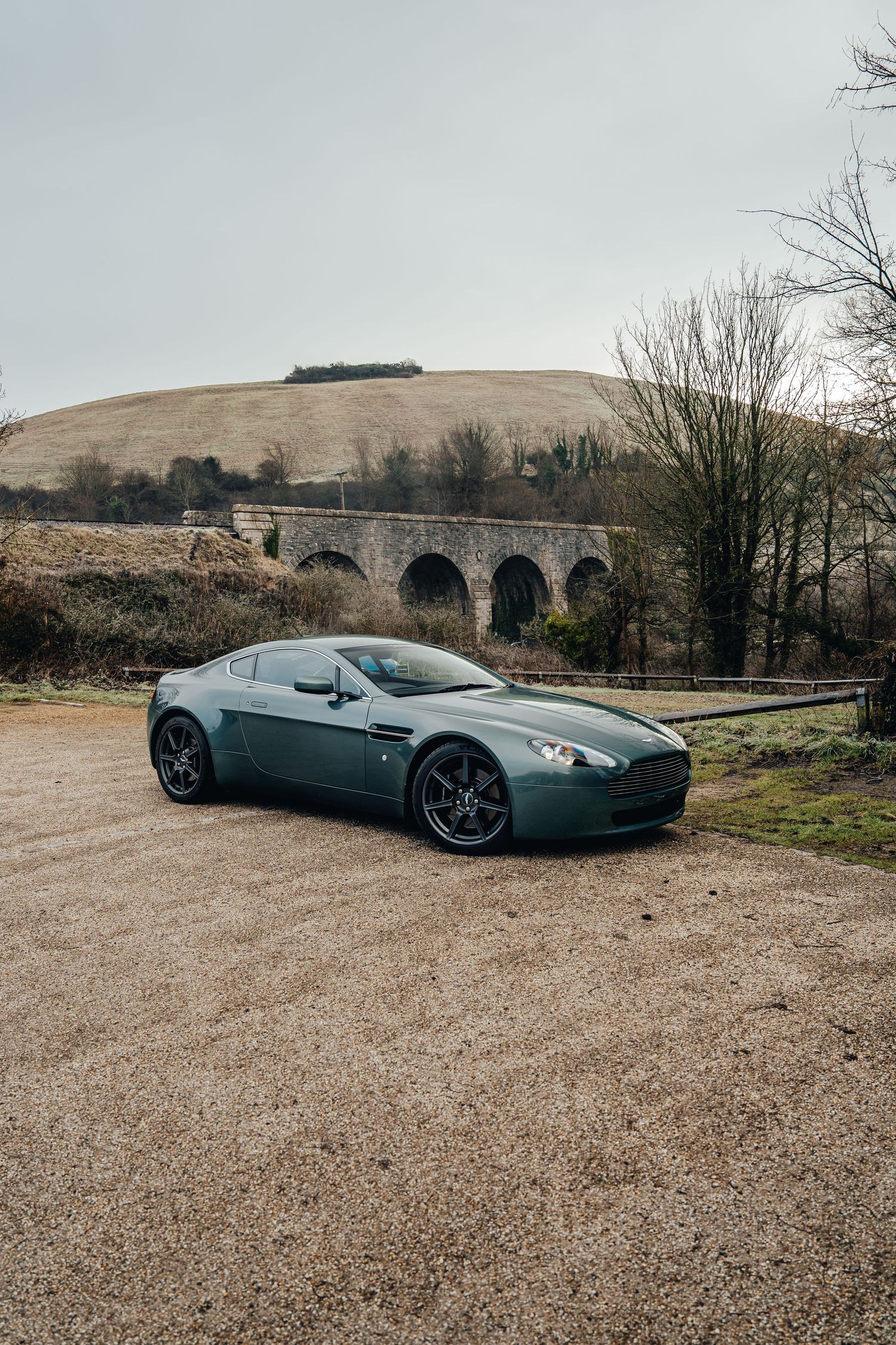 A sleek Aston Martin sports car parked on gravel with a stone railway viaduct and rolling hills in the background.