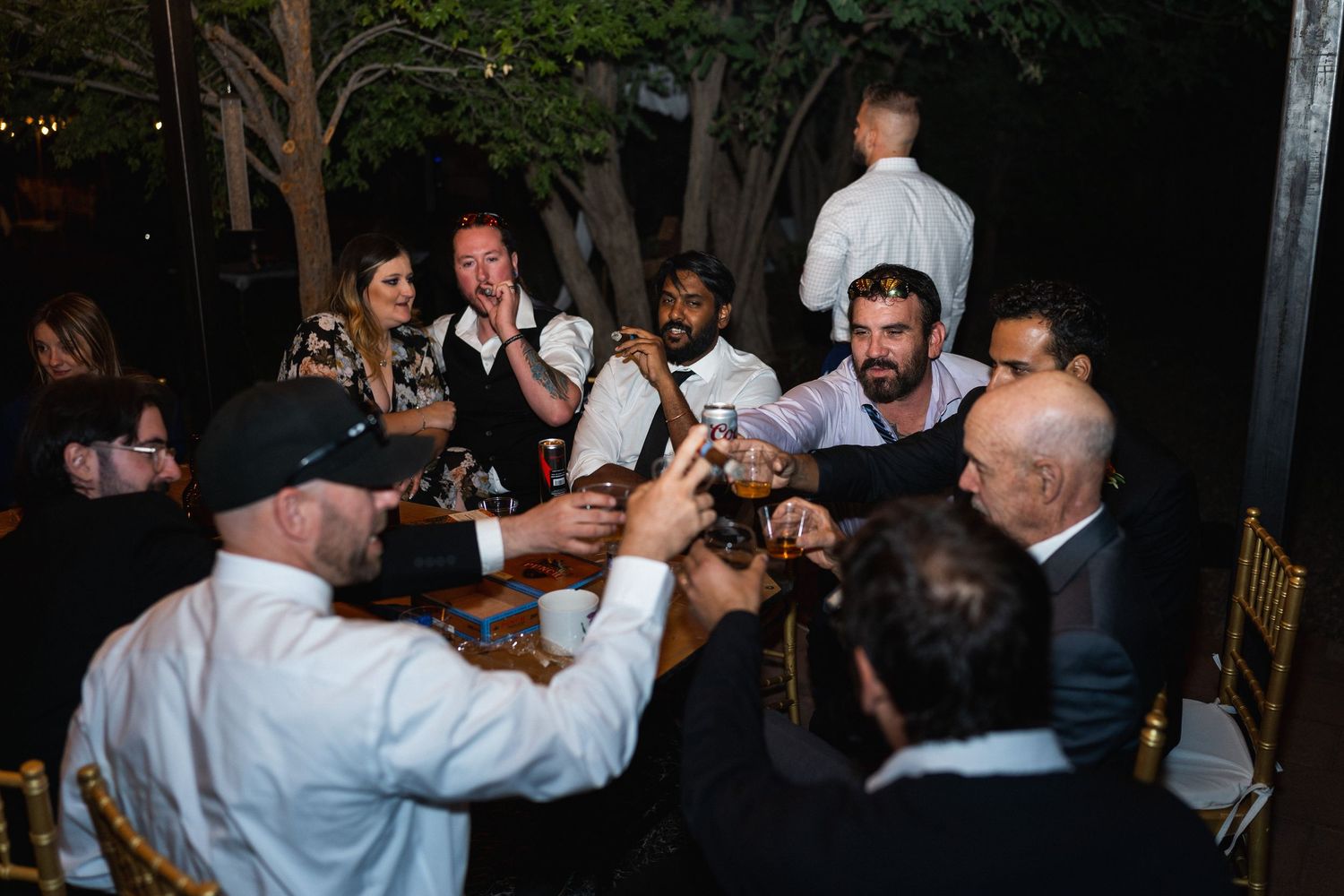 Wedding reception guests raise glasses in a joyful toast around a decorated table at an evening celebration.