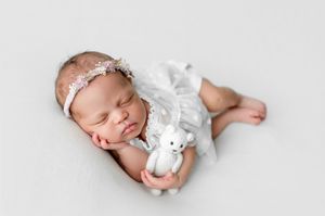 Newborn baby sleeps peacefully in white wrap while holding small stuffed animal toy in studio setting.