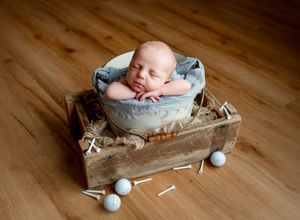 Newborn baby sleeping peacefully in blue bowl on rustic wooden crate with golf balls and tees on hardwood floor.