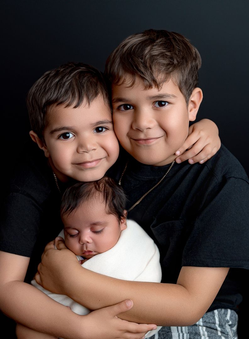 Studio portrait of three siblings in dark clothing against black background, with oldest holding sleeping infant in white wrap.
