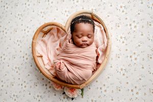 A sleeping newborn baby wrapped in soft pink fabric lies peacefully in a heart-shaped wooden basket.