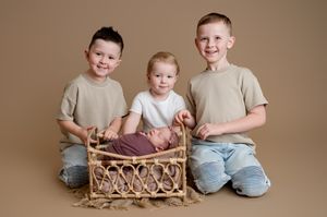 Three siblings pose with a swaddled newborn baby in a wicker basket during a professional studio portrait session.