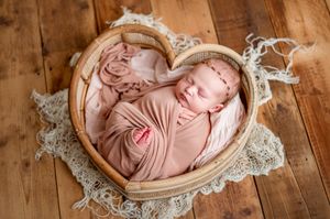 Newborn baby swaddled in pink wrap sleeping in a heart-shaped wooden bowl on a lace blanket.