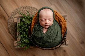 A sleeping newborn wrapped in a green swaddle blanket rests peacefully in a wooden basket.