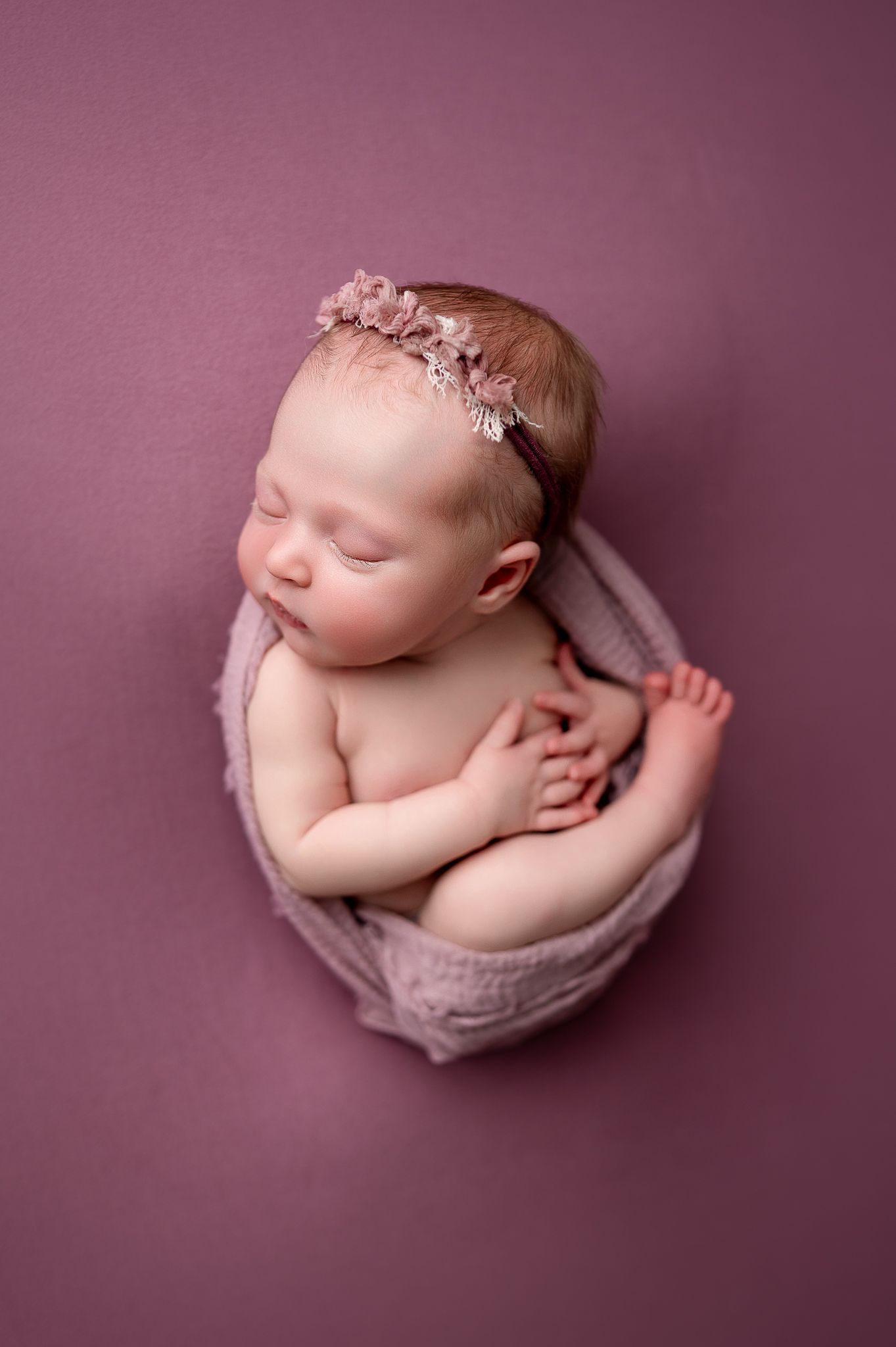Sleeping newborn baby girl in mauve knit bowl wearing pink floral headband on dusty rose background.