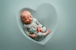 A sleeping newborn baby wearing a light gray onesie lies peacefully on soft mint green fabric.
