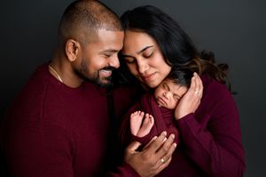 Parents in burgundy sweaters tenderly hold their sleeping newborn baby during a professional studio family portrait session.