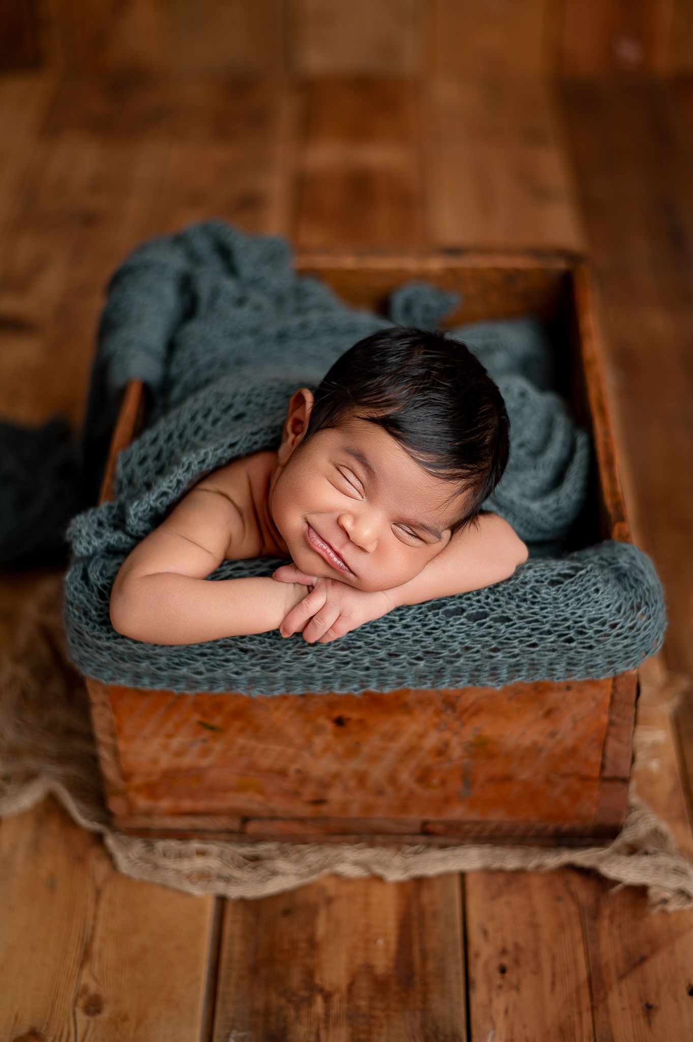 Smiling newborn baby sleeping in a rustic wooden box with teal knit wrap on wood floor.
