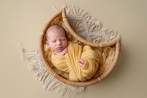 Newborn baby swaddled in yellow wrap sleeping peacefully in a wooden bowl with macrame blanket.