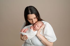 A mother in a white dress tenderly holds her swaddled newborn baby close to her face against a neutral background.