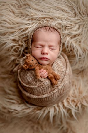 Sleeping newborn baby wrapped in brown knit swaddle with bonnet, holding a knit teddy bear on fluffy beige fur backdrop.
