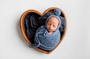 Newborn baby swaddled in blue knit wrap and bonnet, sleeping peacefully in a wooden heart-shaped bowl.