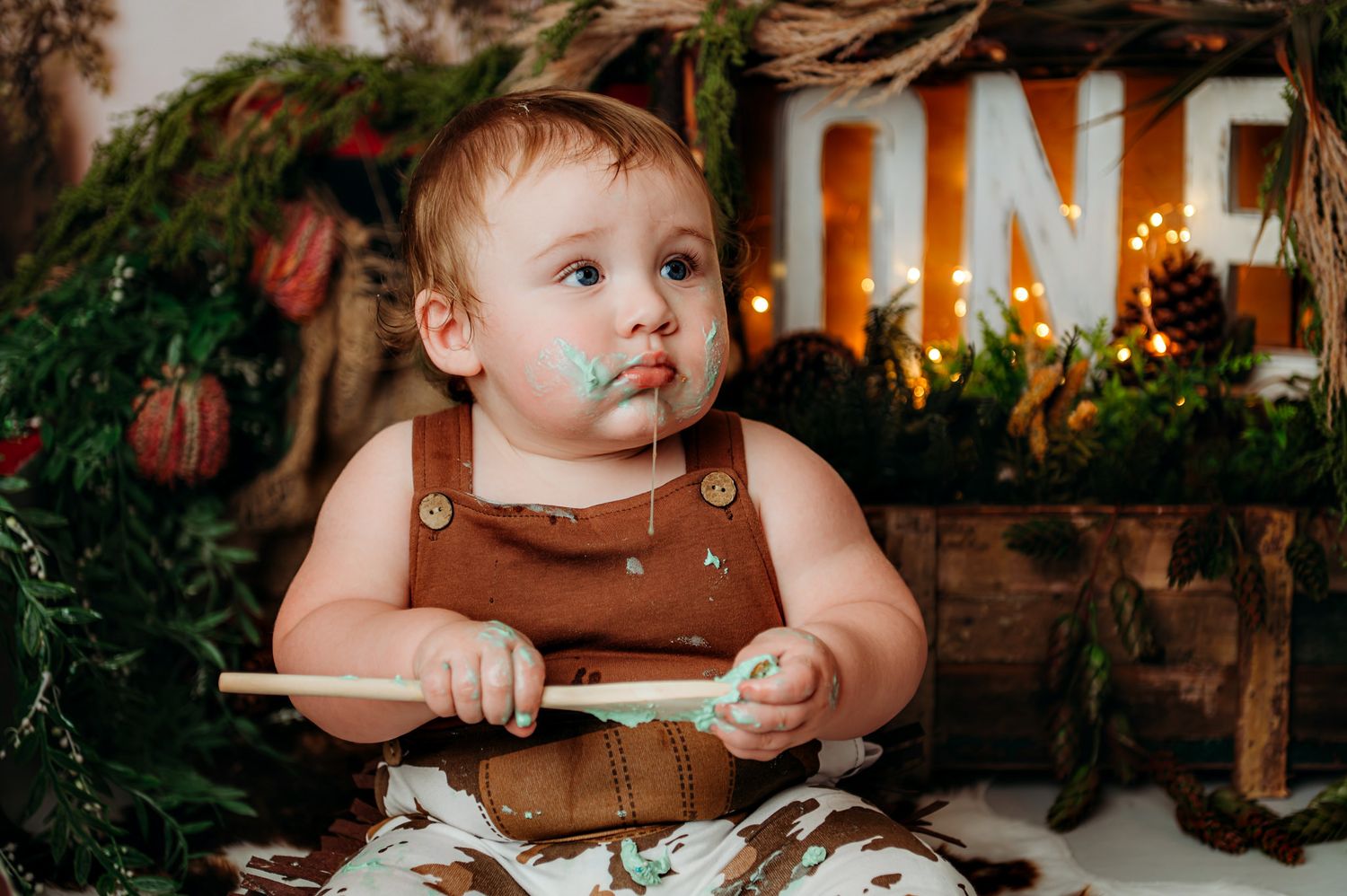 Baby wearing brown overalls sitting outdoors with autumn decor and glowing lights.