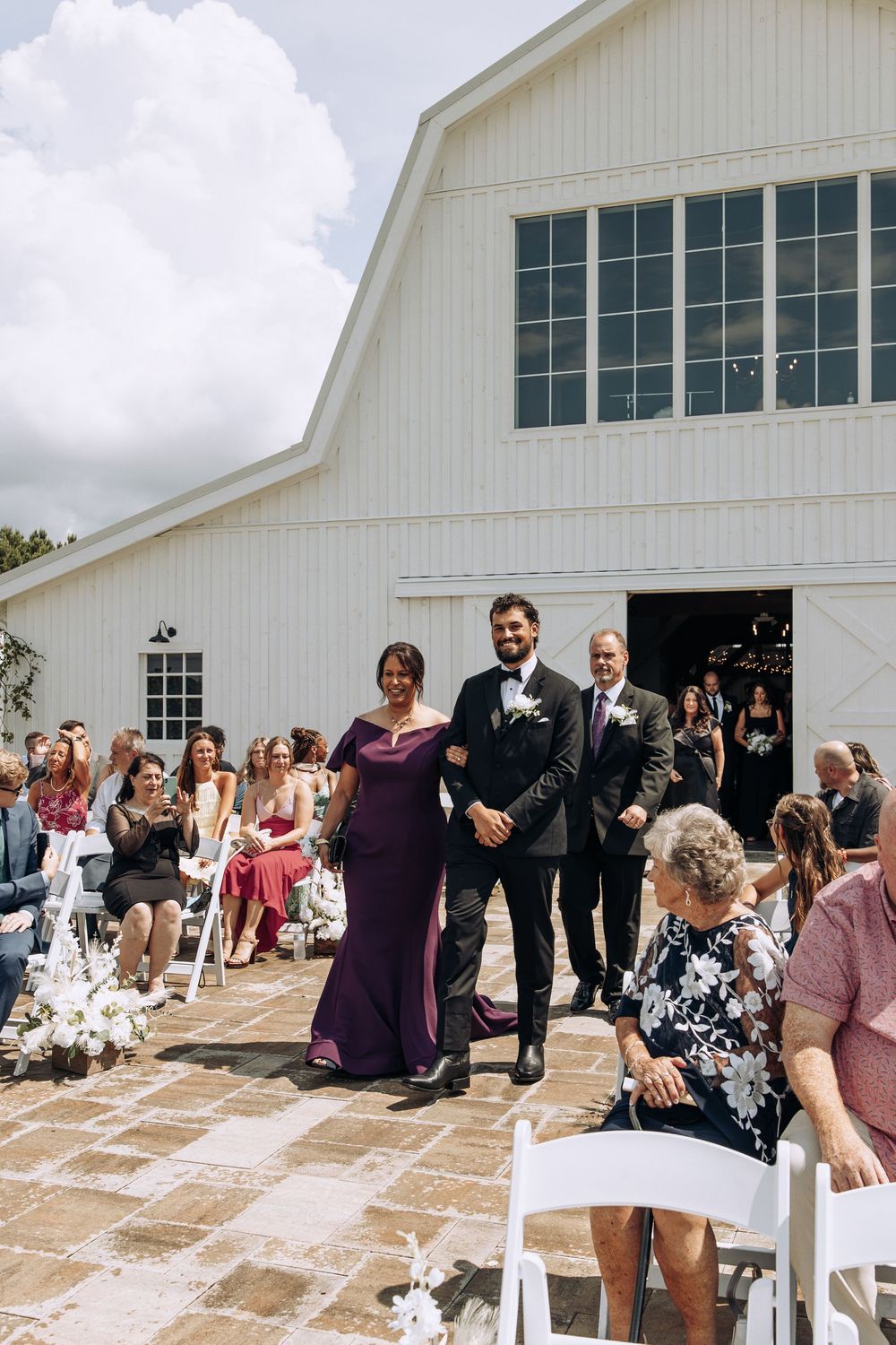 Groom walks down the aisle escorted by parents at outdoor wedding ceremony in front of white barn venue with guests watching.