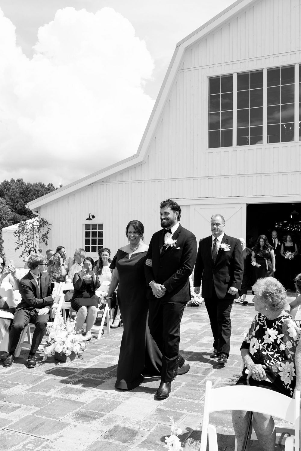 Black and white photo of groom walking down aisle escorted by mother at outdoor wedding ceremony in front of white barn venue.